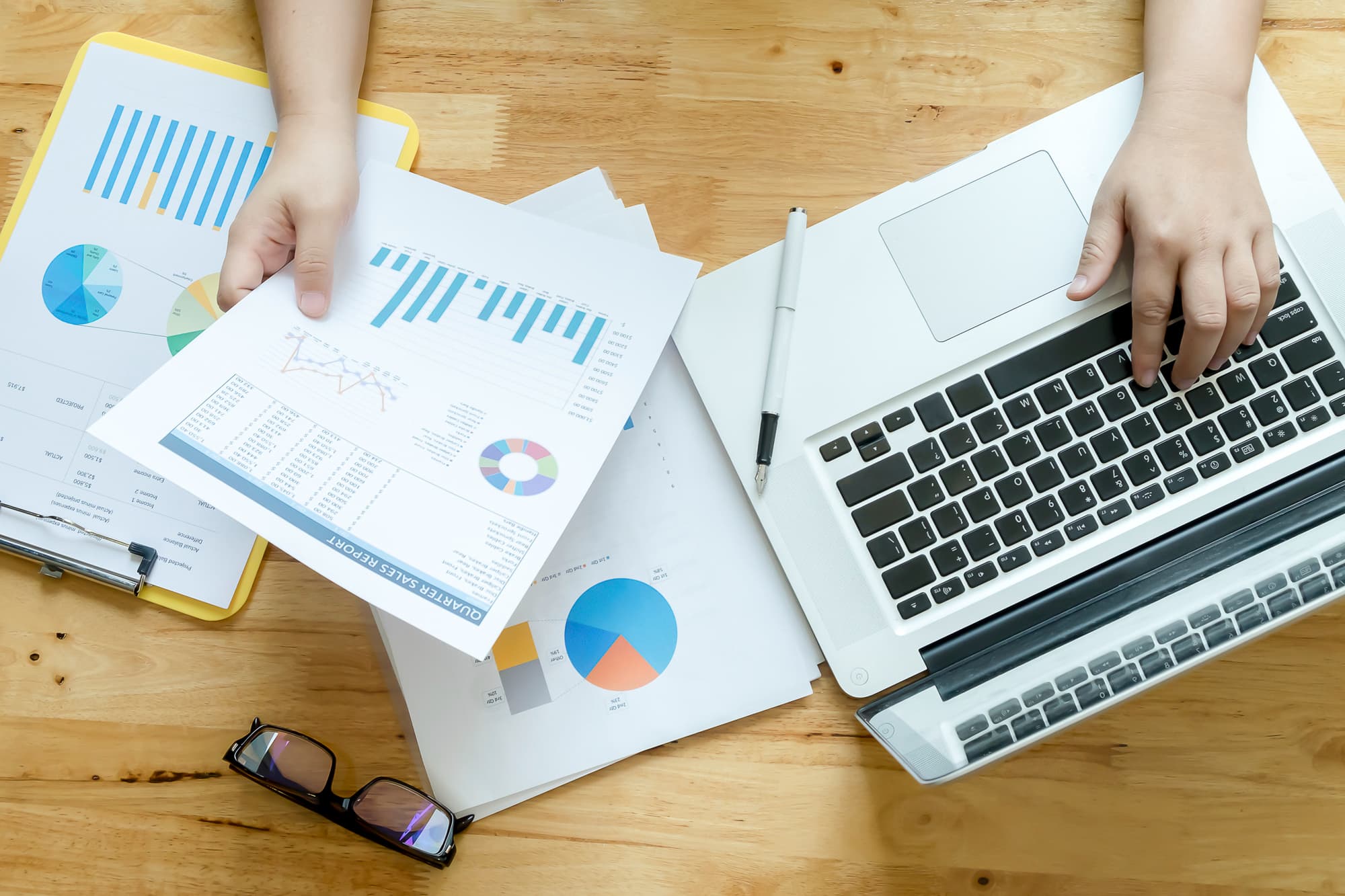 Someone working on a laptop beside printed reports and charts