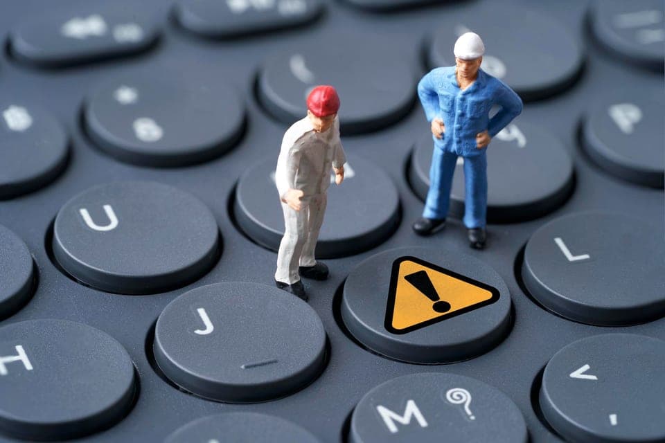 Two worker figurines standing on a keyboard beside a warning sign